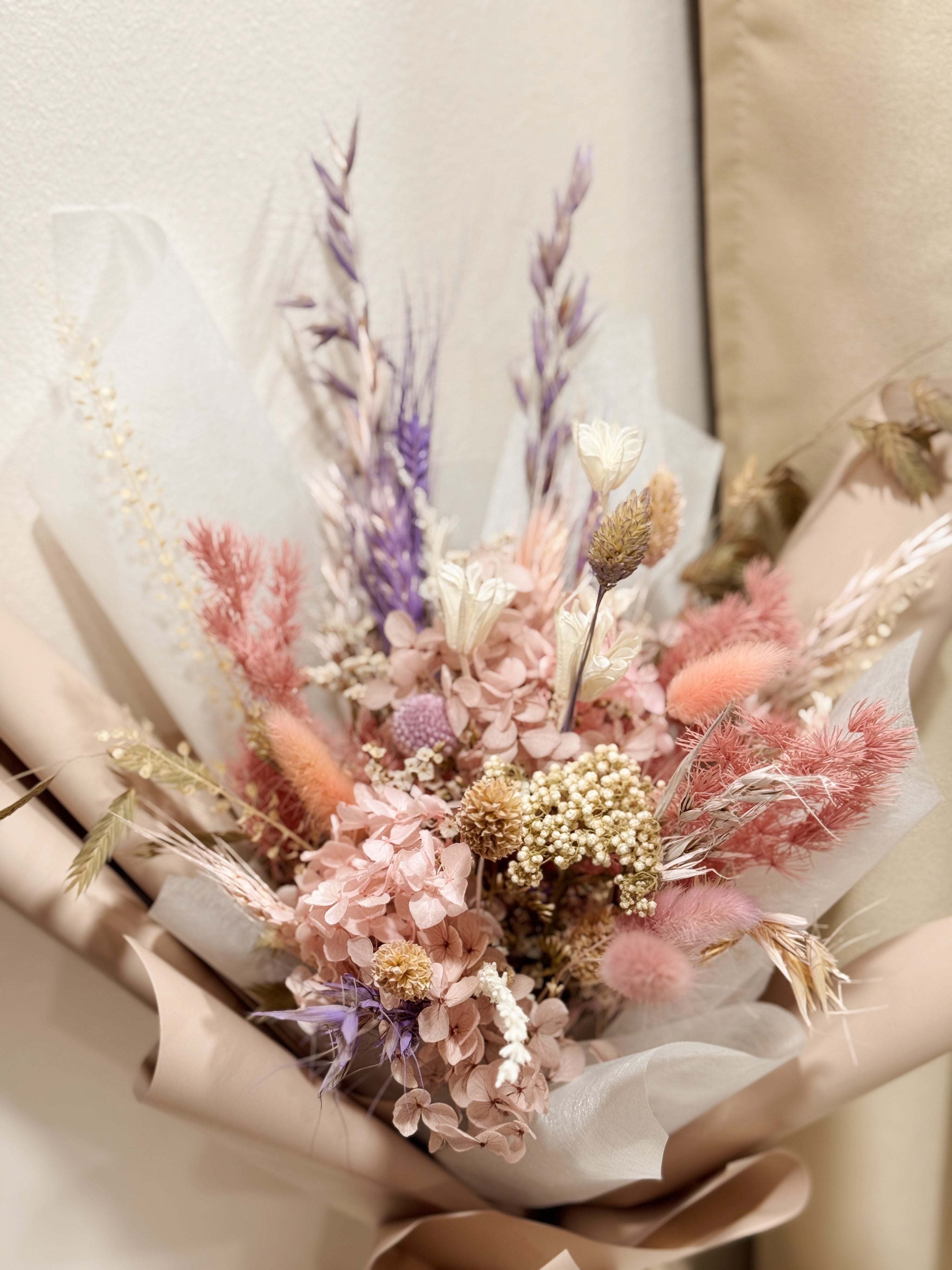 Still Blooming is a delicate preserved flower bouquet featuring soft pink and lavender blooms, thoughtfully gathered with textured foliage. Wrapped in white tissue and blush pink glassine, it is gently crafted by Eden’s Ground, a floral design boutique.