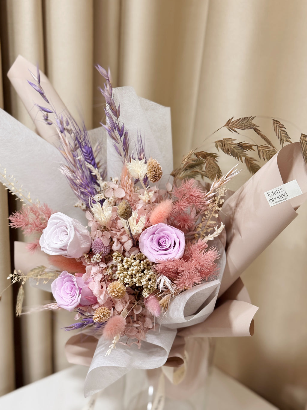 Still Blooming is a delicate preserved flower vase arrangement featuring soft pink and lavender blooms, thoughtfully gathered with textured foliage. Wrapped in white tissue and blush pink glassine, it is gently crafted by Eden’s Ground, a floral design boutique.