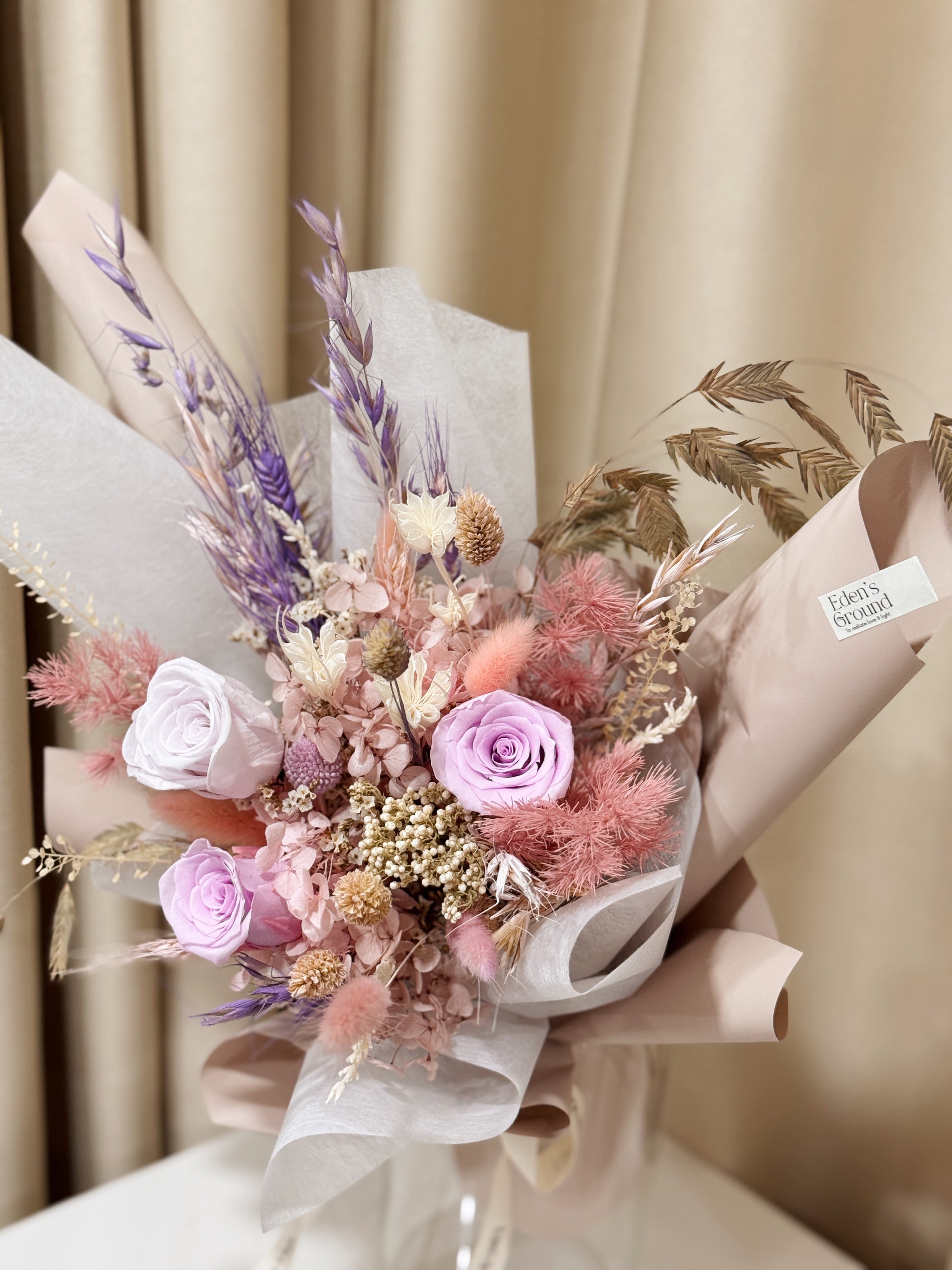 Still Blooming is a delicate preserved flower vase arrangement featuring soft pink and lavender blooms, thoughtfully gathered with textured foliage. Wrapped in white tissue and blush pink glassine, it is gently crafted by Eden’s Ground, a floral design boutique.