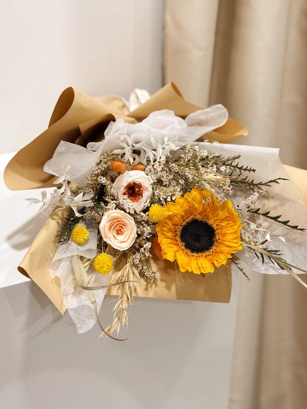 Sunflower Fields Forever bouquet made with preserved sunflower and roses, along with filler flowers and foliage, wrapped in brown kraft paper crafted by Eden’s Ground, a floral design boutique.