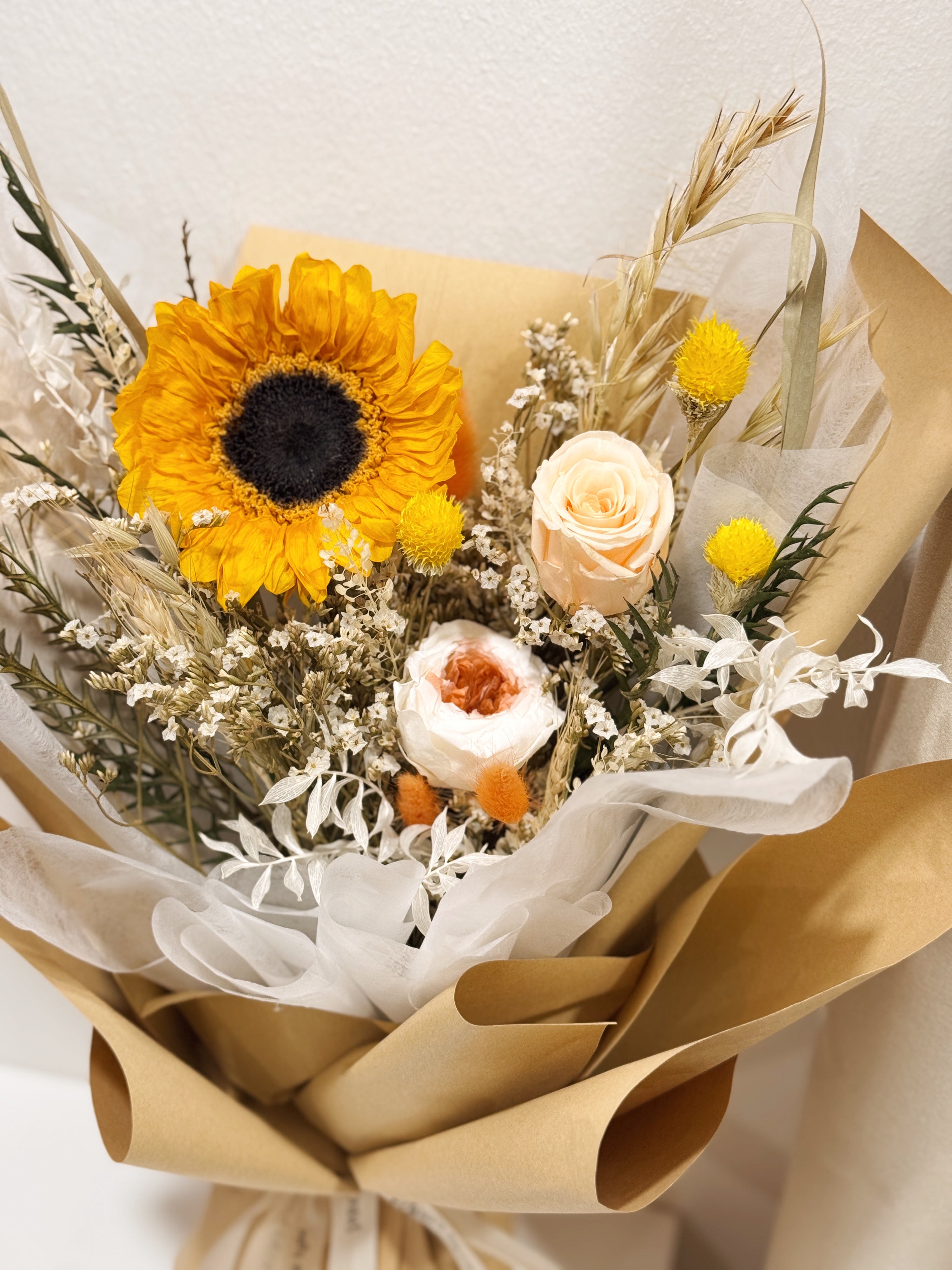 Sunflower Fields Forever bouquet made with preserved sunflower and roses, along with filler flowers and foliage, wrapped in brown kraft paper crafted by Eden’s Ground, a floral design boutique.