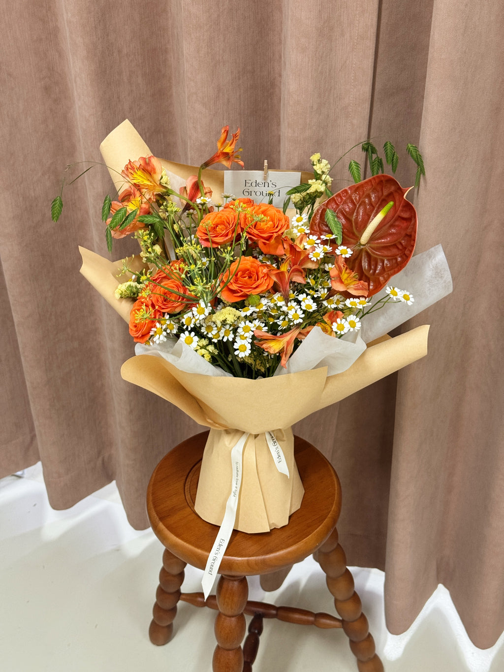 A vibrant floral bouquet featuring orange roses, orange anthuriums, white daisies, and green foliage, wrapped in brown kraft paper and a white ribbon, freshly handcrafted by Eden's Ground, a floral design studio in Singapore.