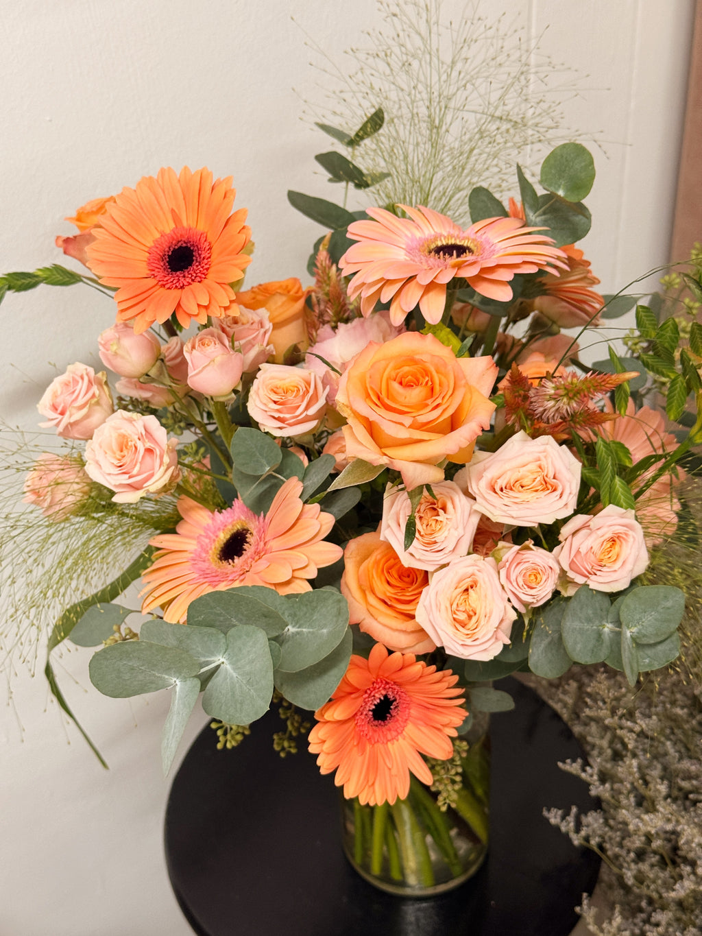 A vibrant floral arrangement featuring orange and peach gerbera daisies, peach roses, and green eucalyptus leaves in a clear glass vase, freshly curated by Eden's Ground a floral design studio in Singapore.