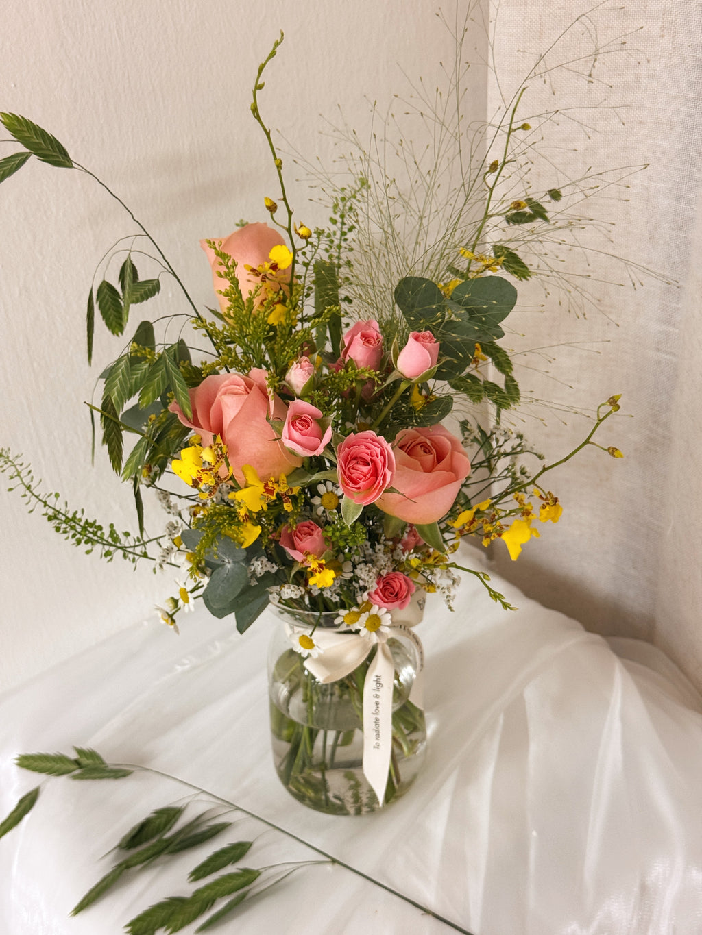 A floral arrangement of pink roses, yellow wildflowers, and green foliage in a clear glass jar vase with a cream ribbon, freshly curated by Eden's Ground, a floral design studio in Singapore.