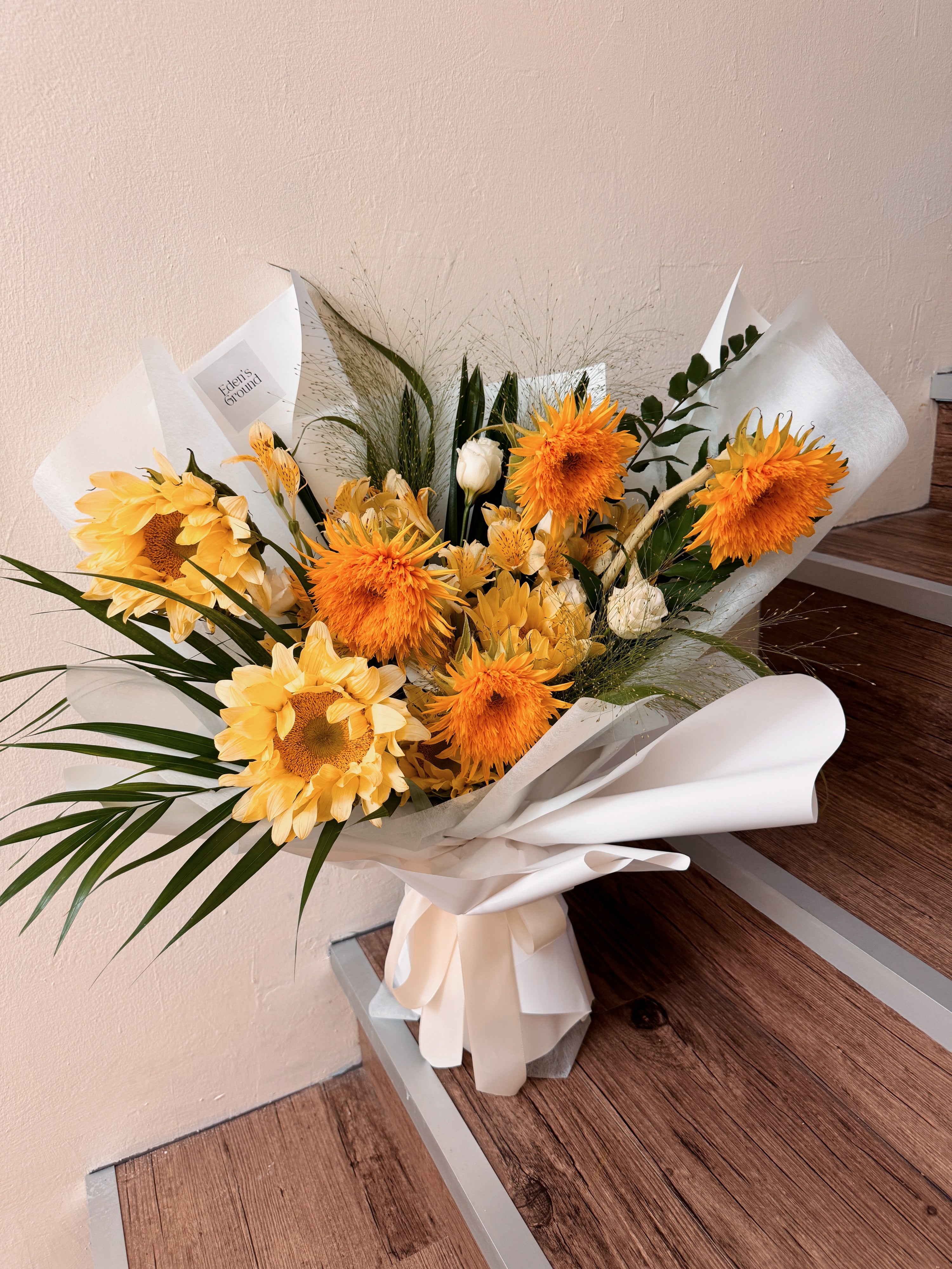 Mix of sunflowers bouquet featuring different types of sunflowers variations, along with filler flowers and foliage like eucalyptus, wrapped in tissue paper and white glassine paper, crafted by Eden's Ground.